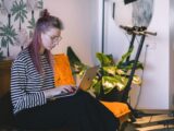 A woman with dyed hair types on a laptop while sitting on a couch in a stylish indoor setting with a scooter nearby.
