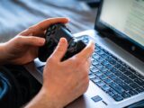 Person holds a gaming controller while interacting with a laptop, emphasizing modern gaming technology.