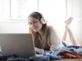 Young woman enjoying music and working on laptop in a cozy bedroom setting.