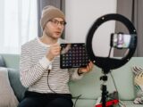 Adult man wearing a beanie records a tech review vlog indoors using a smartphone and ring light.
