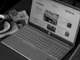 Black and white image of a laptop displaying news articles, accompanied by a cup of coffee and newspapers.