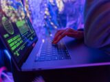 Close-up of hands typing on a laptop displaying cybersecurity graphics, illuminated by purple light.