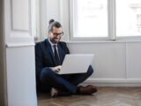 A cheerful man in a suit working remotely on a laptop seated on the floor by a window.