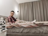 Asian man sitting in bedroom, observing futuristic robot on nightstand.