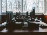 An empty computer lab with multiple workstations and large windows during the daytime.