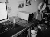 A desk with computers, fan, and storage shelves.