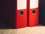 Close-up of two red lever arch files on a wooden desk in a modern office setting.