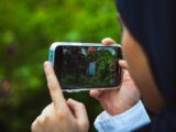 Close-up of person using smartphone to film a green outdoor scene in daylight.