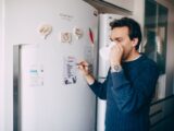 Young male employee in casual outfit drinking coffee and taking notes on paper attached on fridge in morning