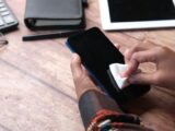 Hands wiping a smartphone with a cloth in an organized workspace. Emphasizes hygiene and device care.