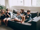 Three children on a gray sofa engrossed in digital devices, indoors.