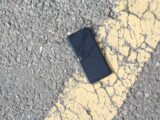 A close-up view of a cracked smartphone lying on a sunlit asphalt road.
