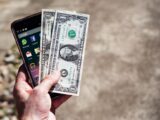 Close-up of a hand holding US dollar bills and a smartphone outdoors, showcasing financial technology.